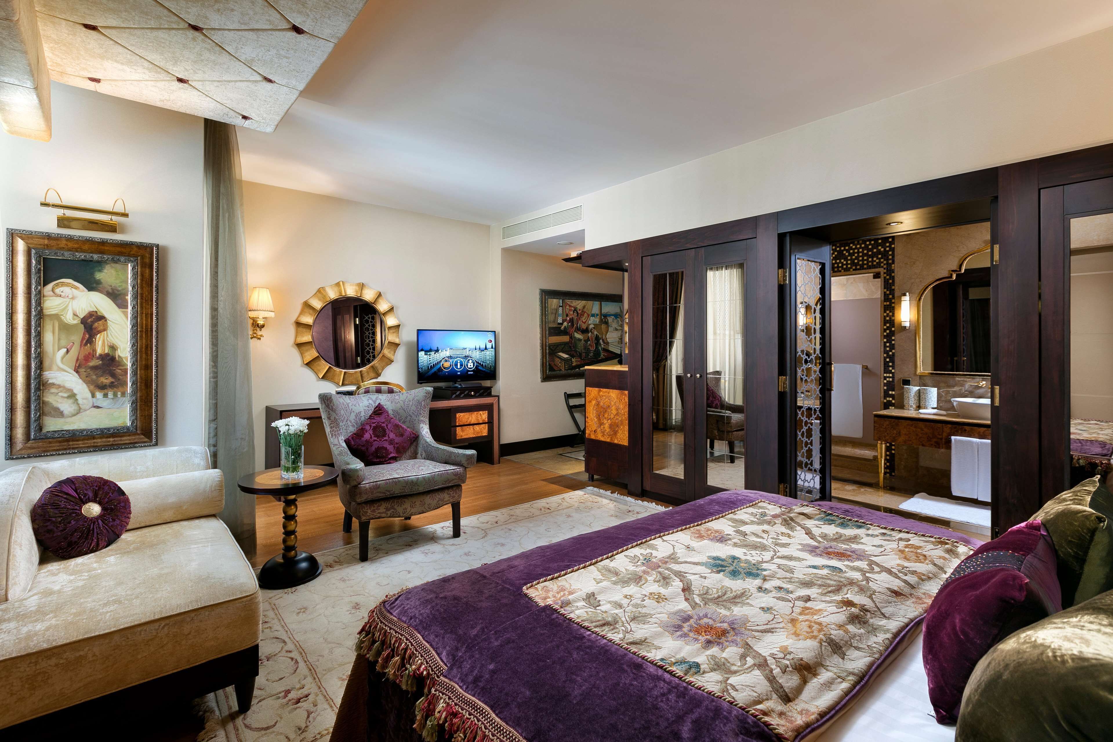A spacious bedroom with a large bed, a sitting area with a sofa and armchair, a television, and a bathroom with a large mirror and sink.