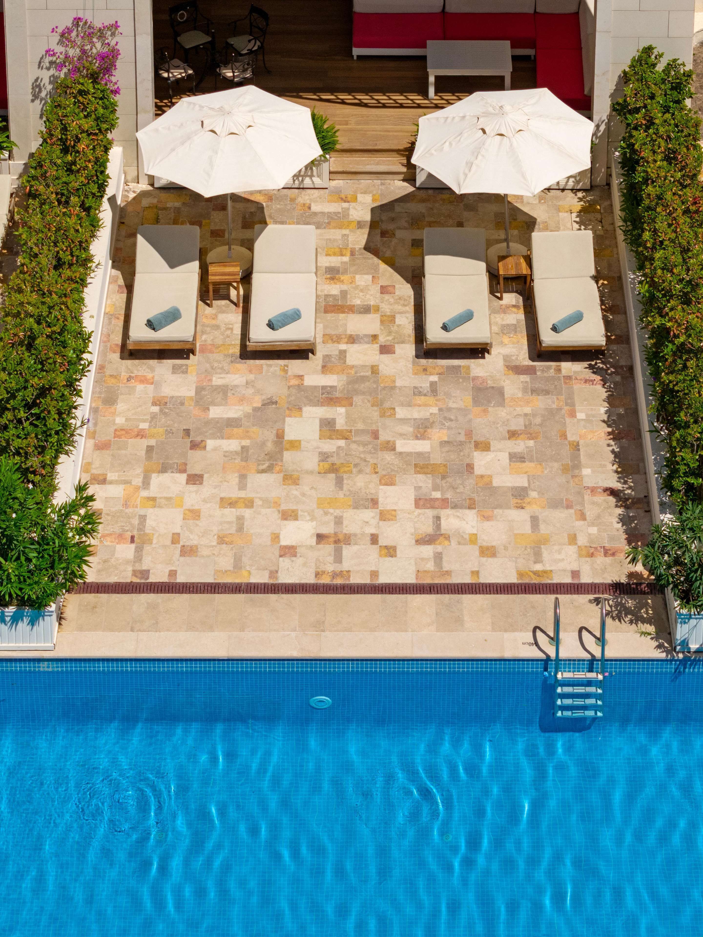 An aerial view of a pool area with four lounge chairs and two umbrellas, surrounded by greenery and a tiled deck.