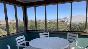 Un solárium con una mesa redonda y sillas blancas, ventanas amplias y vistas a casas y árboles.