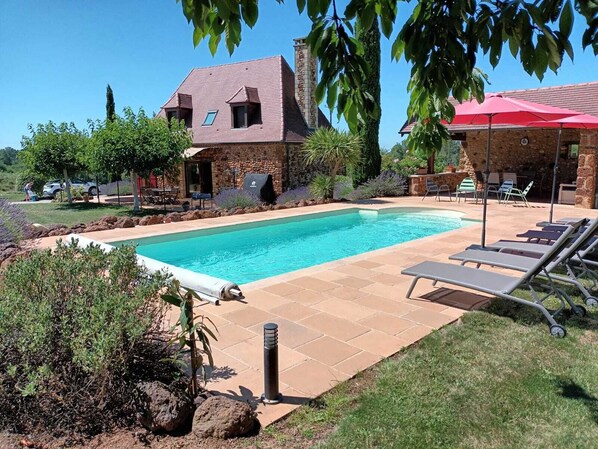 A swimming pool with lounge chairs, a stone house, and a barbecue area.