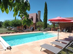 A swimming pool with lounge chairs and a red umbrella, surrounded by stone buildings and greenery.