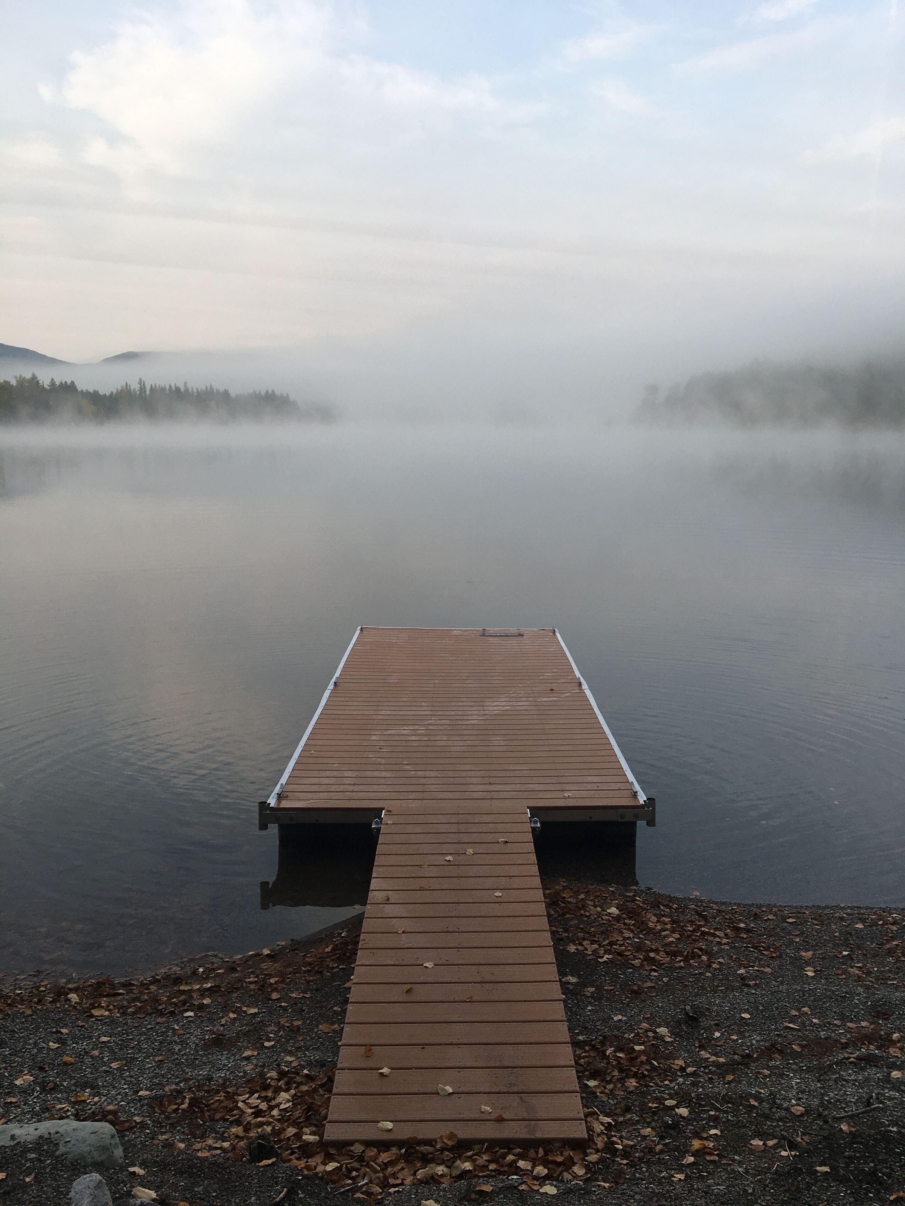 Un muelle de madera se adentra en un lago tranquilo con un bosque brumoso al fondo.