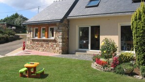 Een huis met een stenen aanbouw, een klein terras met een tafel en stoelen, en een tuin vol kleurrijke bloemen.