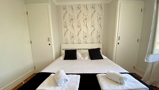 Un dormitorio con una cama grande, ropa de cama blanca, almohadas negras y un papel tapiz floral. Hay dos toallas sobre la cama.