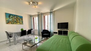 Una sala de estar moderna con un sofá verde, una mesa de centro de vidrio, un comedor con sillas y una mesa, un cuadro en la pared y un televisor sobre un mueble.