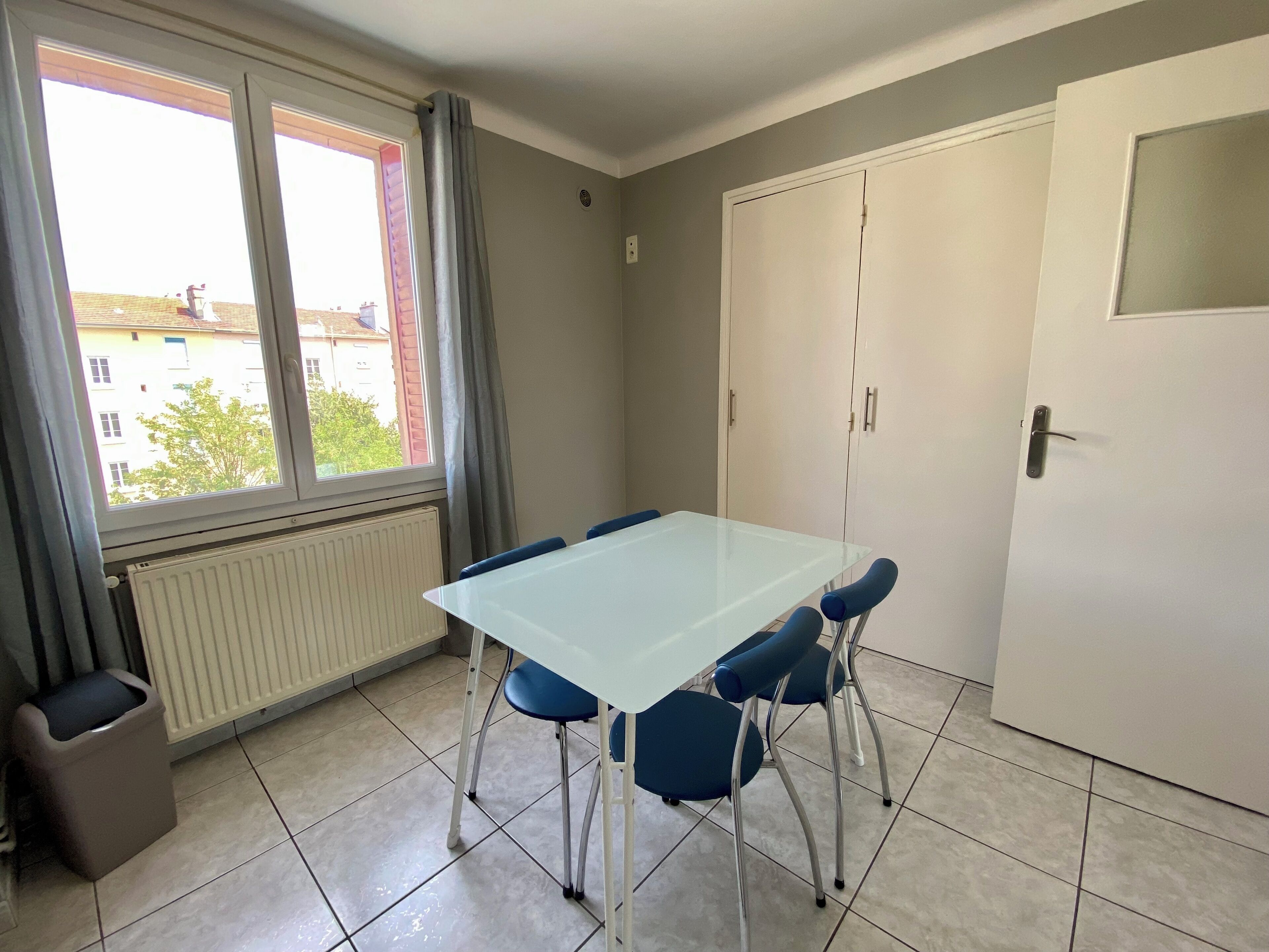 A room with a white table and blue chairs, a radiator, a window with curtains, and a door.