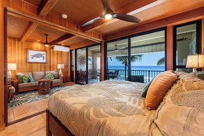 A bedroom with a large bed, a ceiling fan, a wooden ceiling, a window with a view of the ocean, and a balcony with chairs.