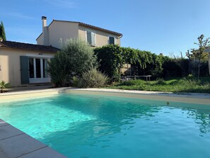 Ein Schwimmbad mit glasklarer, blauer Wasseroberfläche, umgeben von einem Haus mit einem Ziegeldach und einem Garten mit verschiedenen Pflanzen.