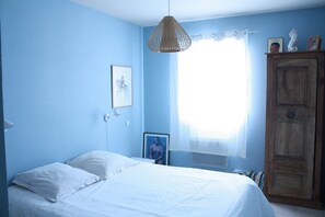 Ein Schlafzimmer mit einem Bett, einem Holzschrank, einer Pendelleuchte und einem Fenster mit transparenten Vorhängen.