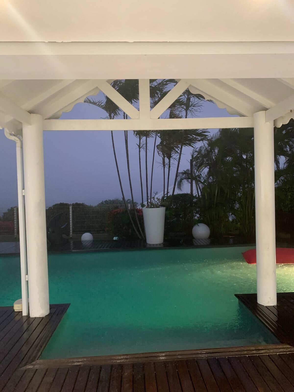 Zona de piscina techada con vista a una piscina azul cristalina, rodeada de palmeras y una cerca.