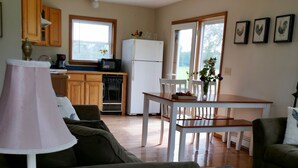 Un salon avec un canapé, une table basse et une lampe. On y trouve également un coin repas avec une table et des chaises. La cuisine est visible au fond, avec des meubles en bois et un réfrigérateur.