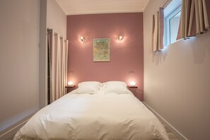 A neatly made bed in a room with a pink wall, two bedside lamps, and a window with curtains.