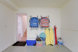 Una habitación con una puerta que da a una escalera, un perchero con objetos doblados, un enfriador azul, un recipiente de plástico, una tabla de surf y un cilindro de gas.