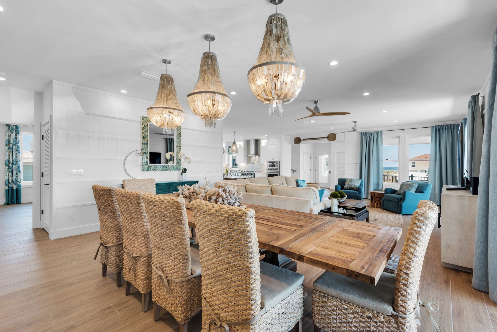 A spacious living area with a large wooden dining table surrounded by wicker chairs, a sofa, and a blue armchair. There are three hanging chandeliers and a ceiling fan.