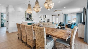 A spacious living area with a large wooden dining table surrounded by wicker chairs, a sofa, and a blue armchair. There are three hanging chandeliers and a ceiling fan. - 🌊 Sealation! – Luxe 8BR w/ Pool | Game Room | Rooftop Deck | Elevator 🌊 (Pensacola Beach)