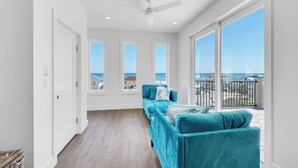 A modern living room with a teal sofa, a ceiling fan, and large windows offering a view of the sea. - 🌊 Sealation! – Luxe 8BR w/ Pool | Game Room | Rooftop Deck | Elevator 🌊 (Pensacola Beach)