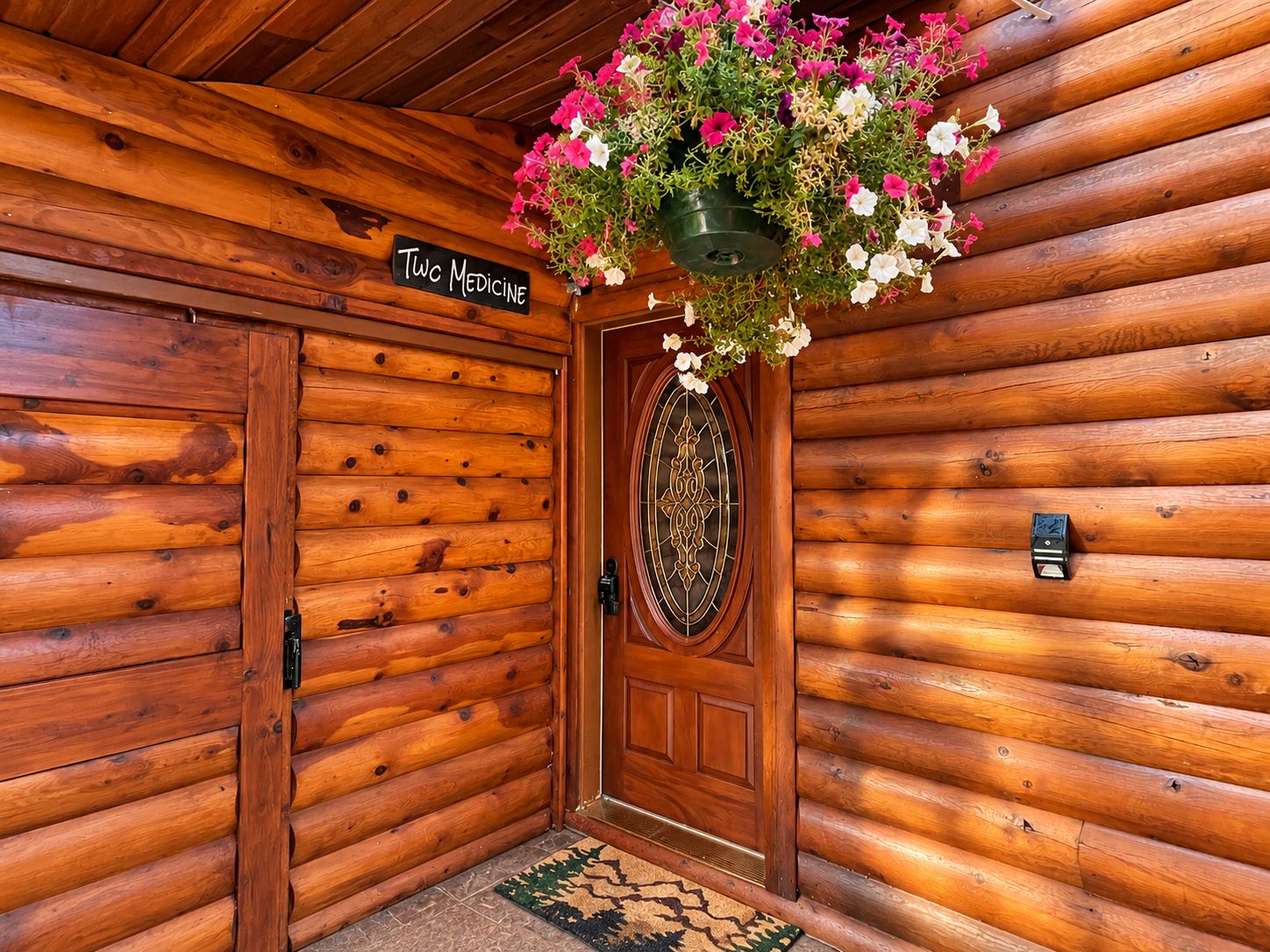 A wooden cabin entrance with a sign reading 'Two Medicine' and a hanging flower basket.