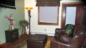 Un salon comprenant un canapé, un fauteuil, un pouf, une lampe sur pied, un téléviseur et une fenêtre avec des stores.