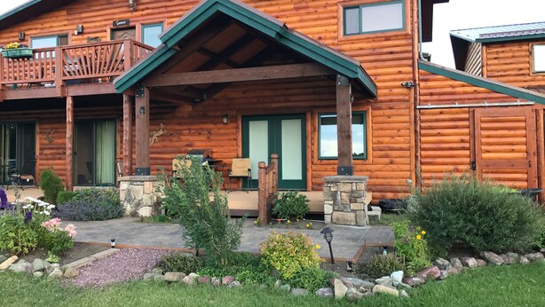 Une cabane en rondins dotée d’un porche couvert, de piliers en pierre et d’un jardin aménagé.