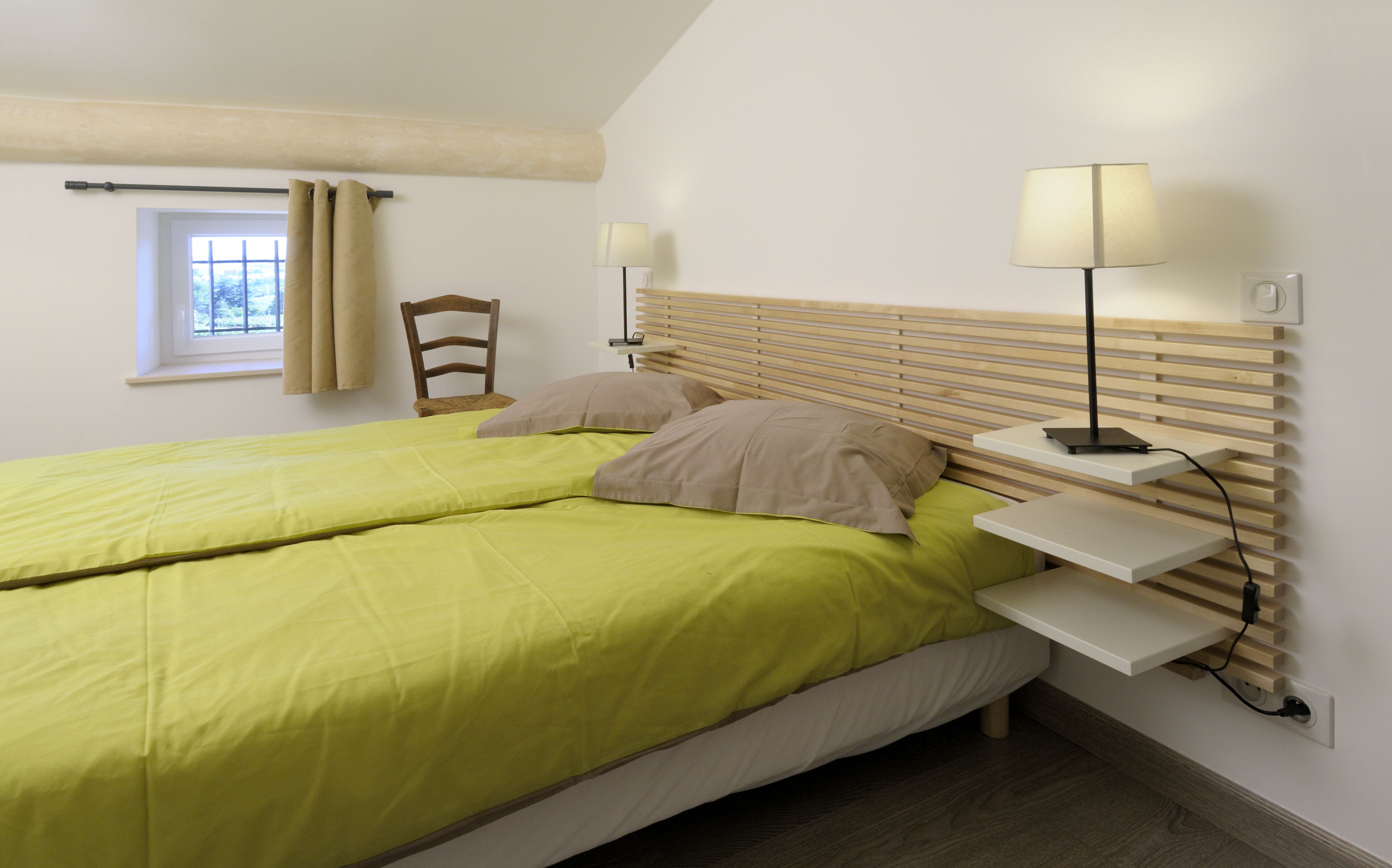 A bedroom with a bed, two bedside tables, a chair, a window with curtains, and a lamp.