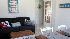 Sala de estar com um sofá escuro, mesa de centro de madeira e cadeiras brancas. Há uma porta com painéis de vidro jateado e um enfeite de peixe fixado na parede.