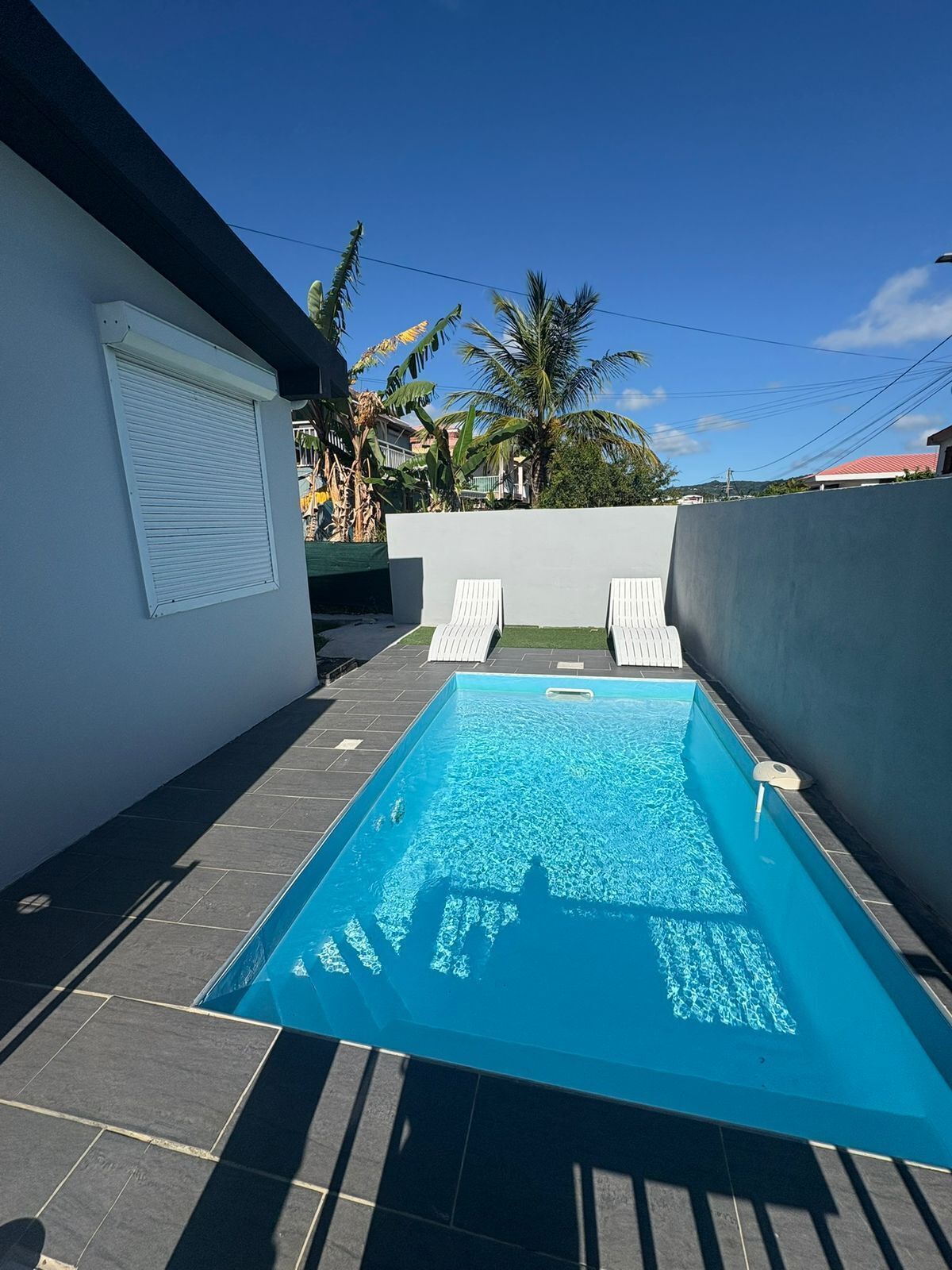 A rectangular swimming pool with two lounge chairs, surrounded by a low wall and a white building with a closed window.