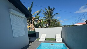 Une piscine rectangulaire avec deux chaises longues, entourée d’un petit muret et d’un bâtiment blanc dont la fenêtre est fermée.