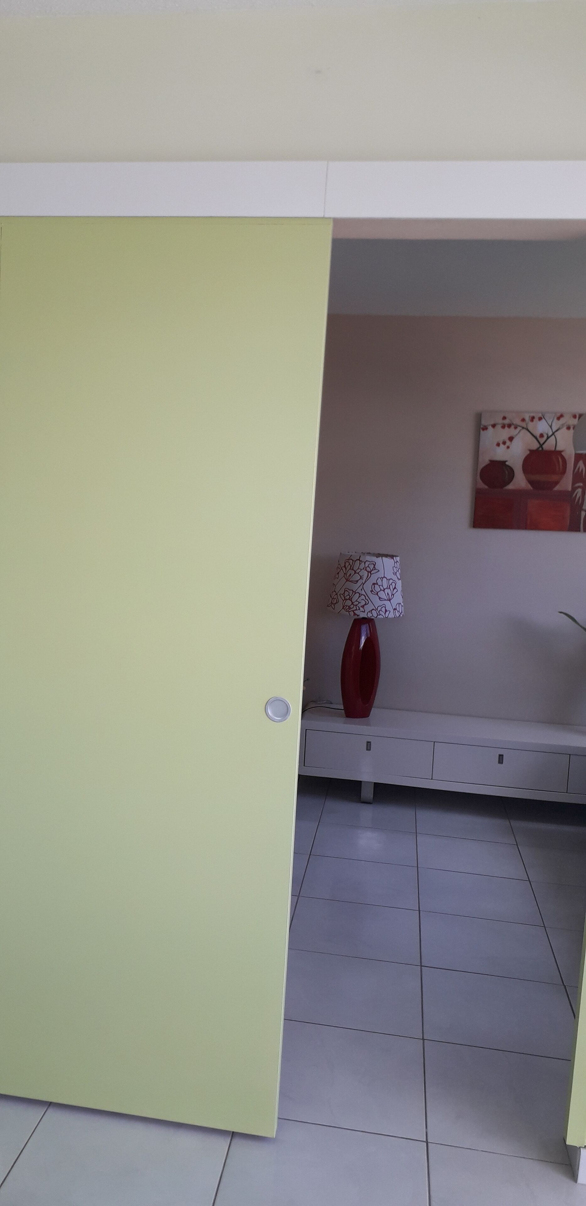 Une pièce avec une porte fermée, un vase rouge posé sur une table blanche et un tableau accroché au mur.