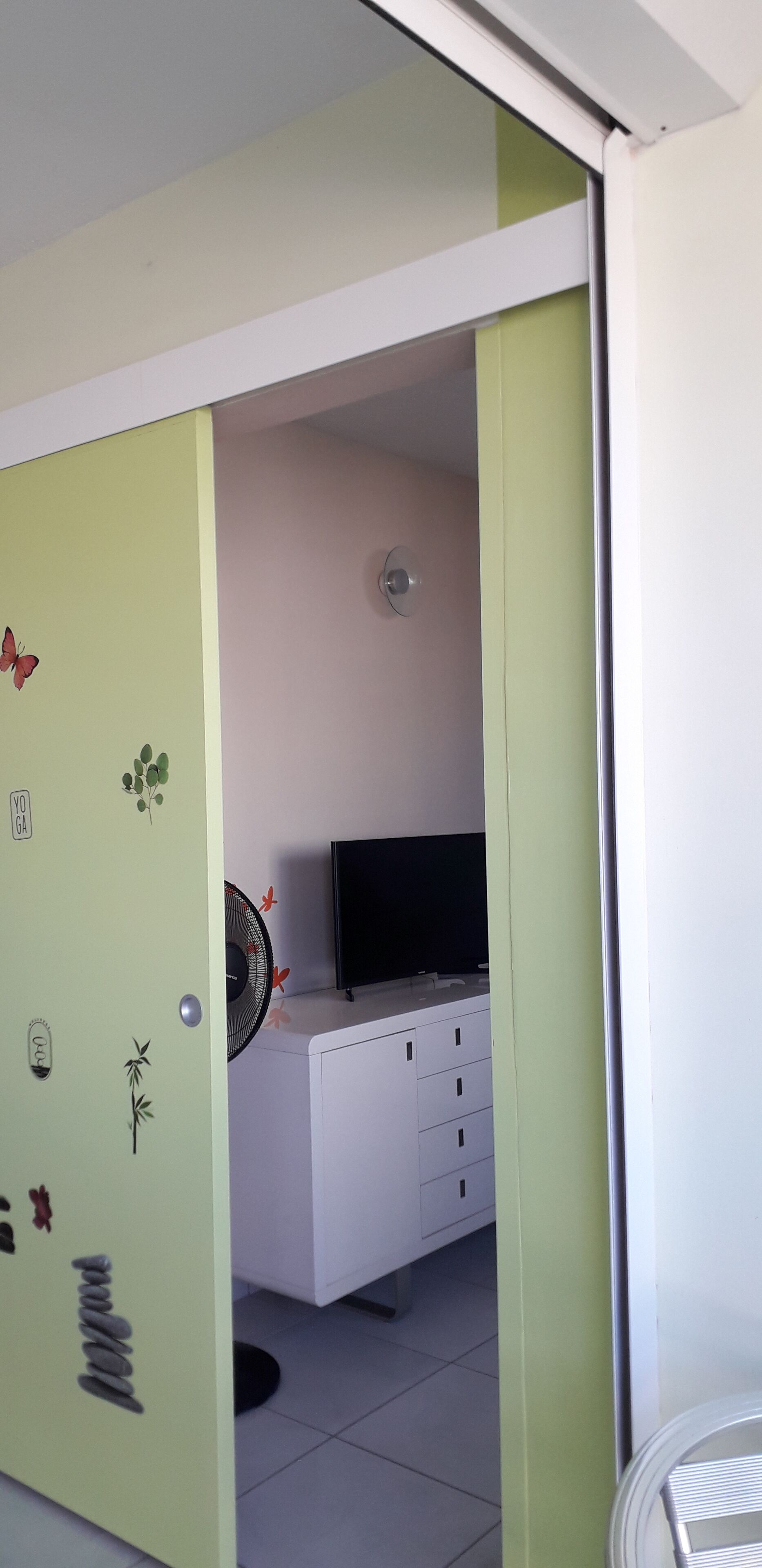 A room with a green door, a white cabinet with drawers, a black television on top, and a fan with red decorations.