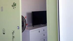 Ett rum med en grön dörr, ett vitt skåp med lådor, en svart tv ovanpå och en fläkt med röda detaljer.