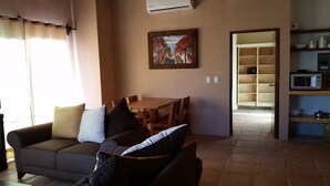 A living room with a sofa, a dining table with chairs, a painting on the wall, and a kitchen area with shelves and a microwave. - 1 Bedroom Apartment in Flat Area Near the Beach and Zocalo  (Bucerías)