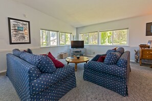 A living room with two patterned sofas, a coffee table, a TV on a stand, and large windows with a view of trees. - Secluded location, private Beach Access, water views, sleeps 6 (Port Angeles)