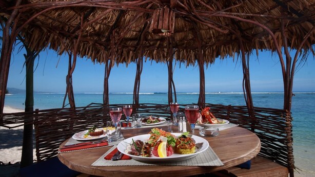 Zona pranzo in spiaggia con tetto di paglia, tavolo in legno e vista sull'oceano.