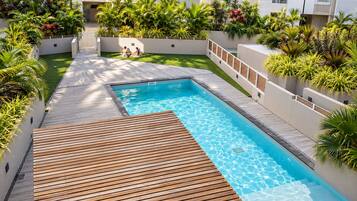 A swimming pool with a wooden deck, surrounded by lush greenery and a white building.