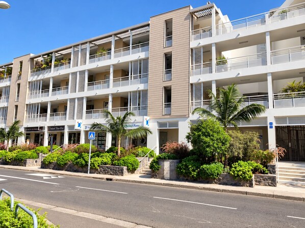 A multi-story hotel with balconies, a street in front, and greenery.