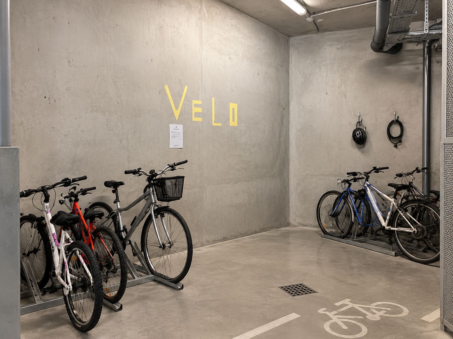 A bicycle parking area with the word 'VELO' on the wall, a bike rack, and a helmet hanging on the wall.