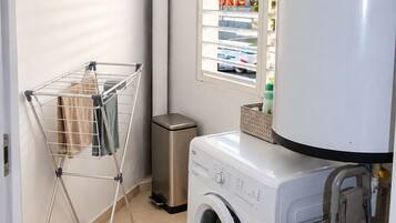 A laundry area with a washing machine, a drying rack, a trash bin, and a window with blinds.