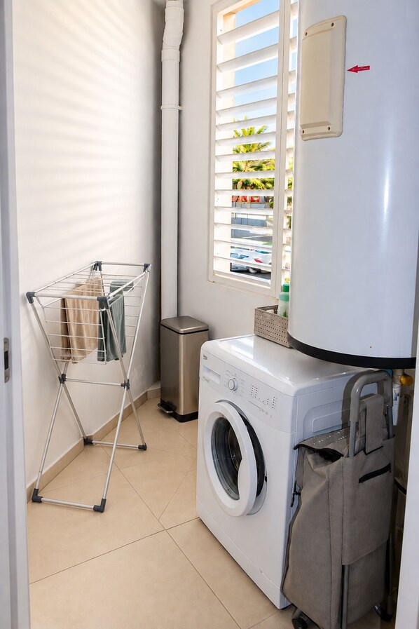 Un espace buanderie comprenant un lave-linge, un étendoir, une poubelle et une fenêtre avec des stores.