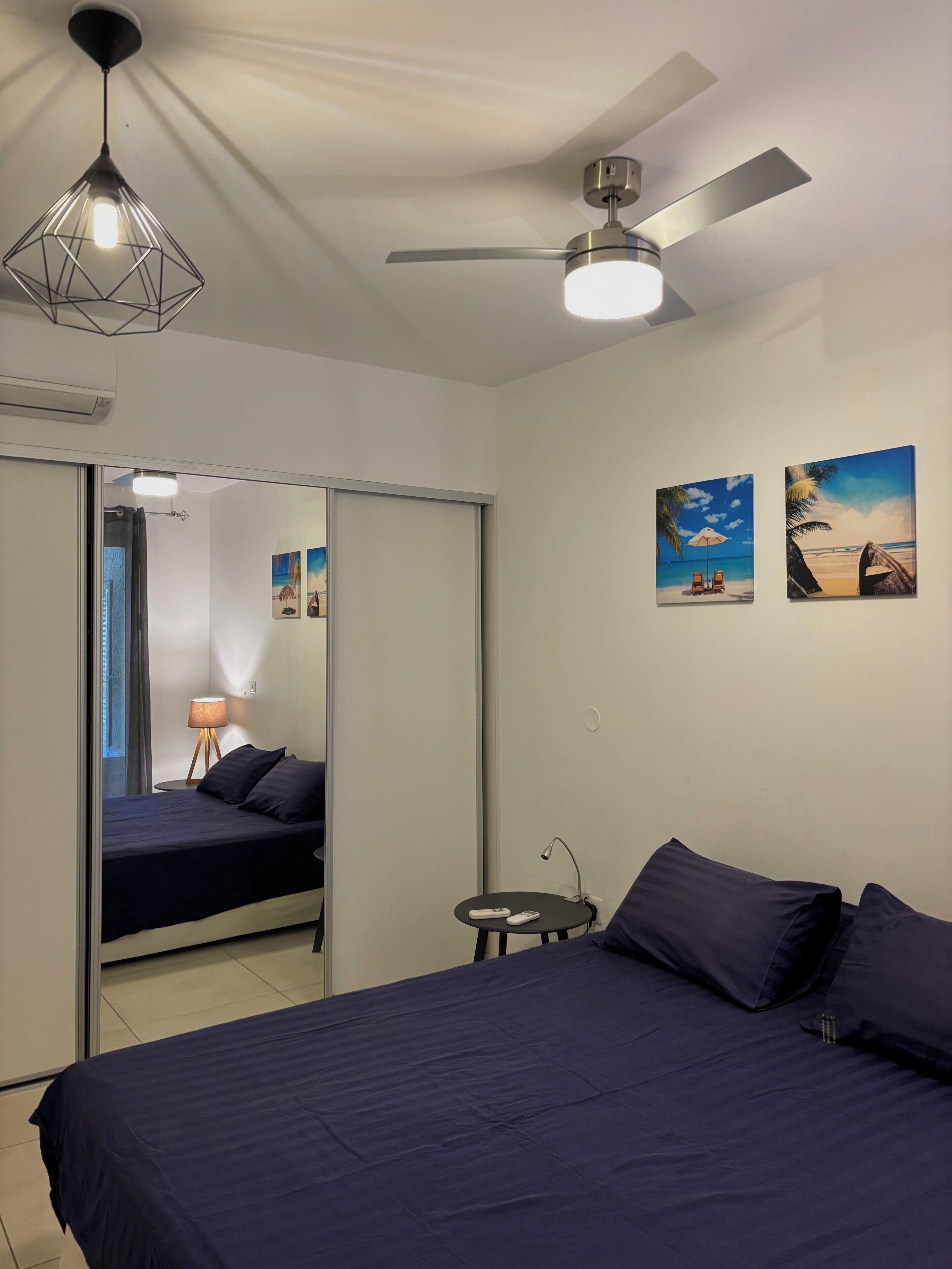 A bedroom with a bed, a ceiling fan, a wall-mounted lamp, and two beach-themed paintings.