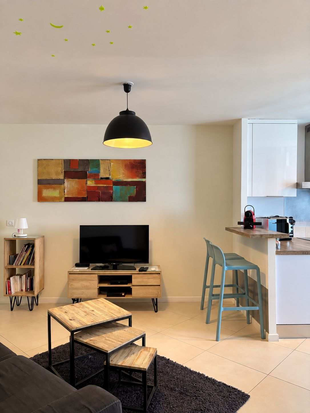 Een moderne woonkamer met een flatscreen televisie, een houten salontafel, een zwarte bank, een blauwe fauteuil en een keuken met witte kastjes.