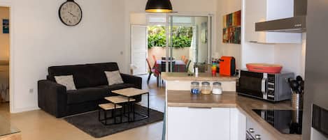 Une cuisine moderne avec un îlot central, un coin repas avec une table et des chaises, un canapé noir agrémenté de coussins à motifs et une horloge murale.