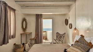 A bedroom with a large bed, a chair, a small table, and a view of the sea through an open door.