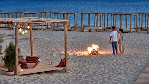 A beach setting with wooden structures and seating areas, a fire pit, and a view of the sea.