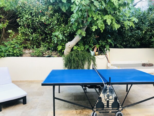Une table de ping-pong se trouve dans un espace patio extérieur, doté d’un sol en pierre, entouré d’une végétation luxuriante et bordé d’un mur blanc.