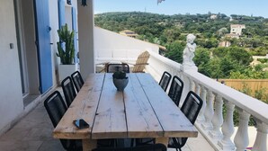Une table en bois, entourée de chaises, est placée sur un balcon, d’où l’on peut admirer un paysage verdoyant et luxuriant, avec des bâtiments visibles au loin.