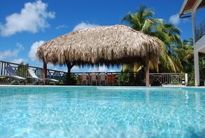 A pool with a thatched roof structure, lounge chairs, and palm trees in the background. - 76m2 apartment on one level, large infinity pool, sea view, garden (Le Robert)