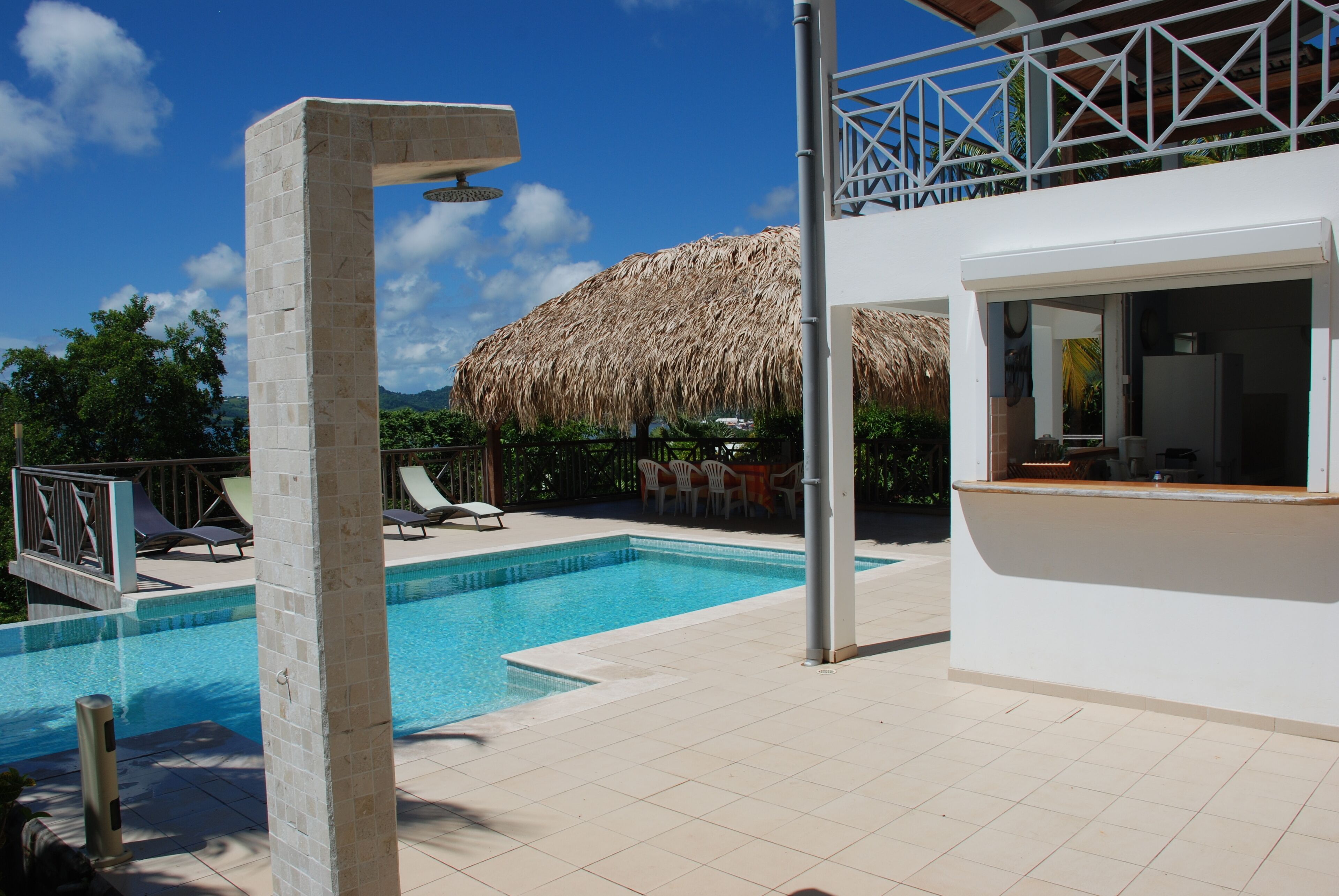 A swimming pool with lounge chairs, a thatched roof structure, and a white building with a balcony.
