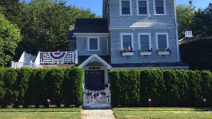 A two-story house with a white picket fence, a small porch, and a flag on the roof. - Experience Hy-View Hyannisport Exceptional Summer Getaway (Hyannisport)