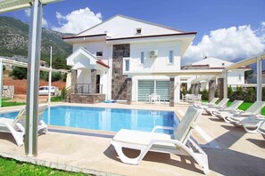A modern villa with a swimming pool, outdoor seating, and a mountain backdrop. - Villa Mendos (Fethiye)