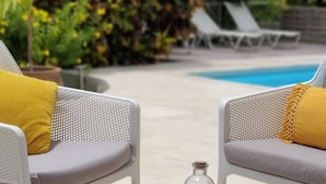 Een zwembadgedeelte met twee witte wicker stoelen, elk met een geel kussen. Op de voorgrond staat een klein tafeltje met een glazen fles en een decoratieve doos.